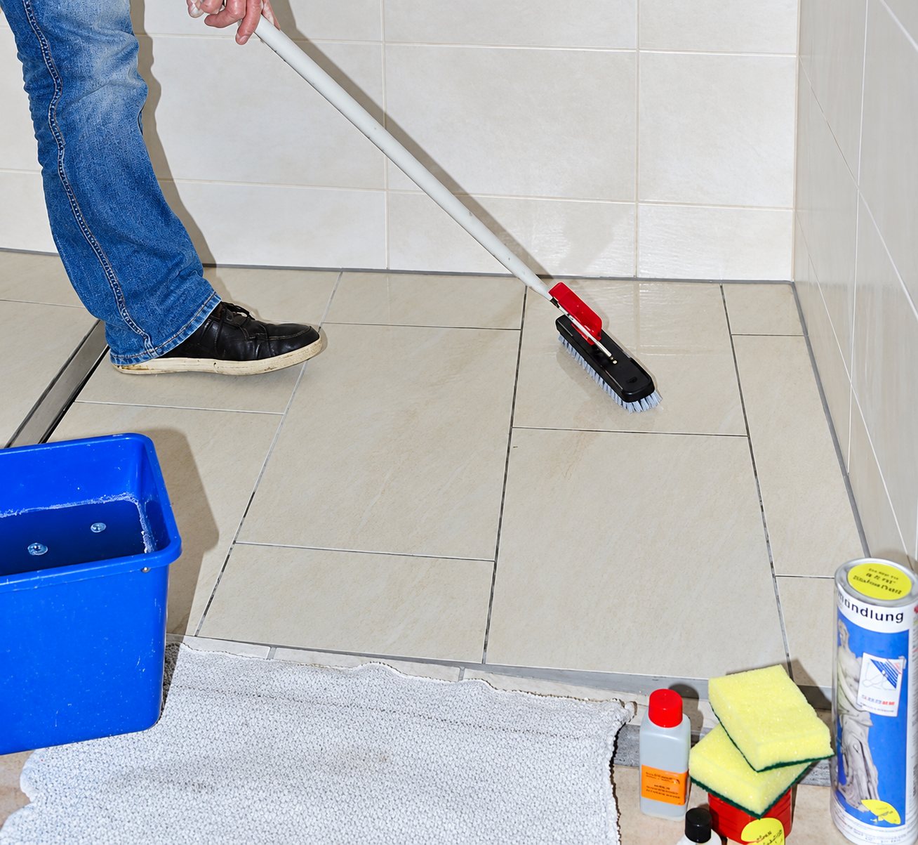 Person in Jeans schrubbt den beigen Fliesenboden einer bodengleichen Dusche, davor Eimer, Schwämme und Pflegemittel