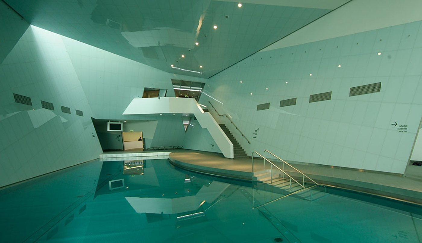 Moderne Schwimmhalle mit schräg verlaufenden, hell gefliesten Wänden, türkisfarbenem Becken und einer Treppe mit Edelstahlgeländer