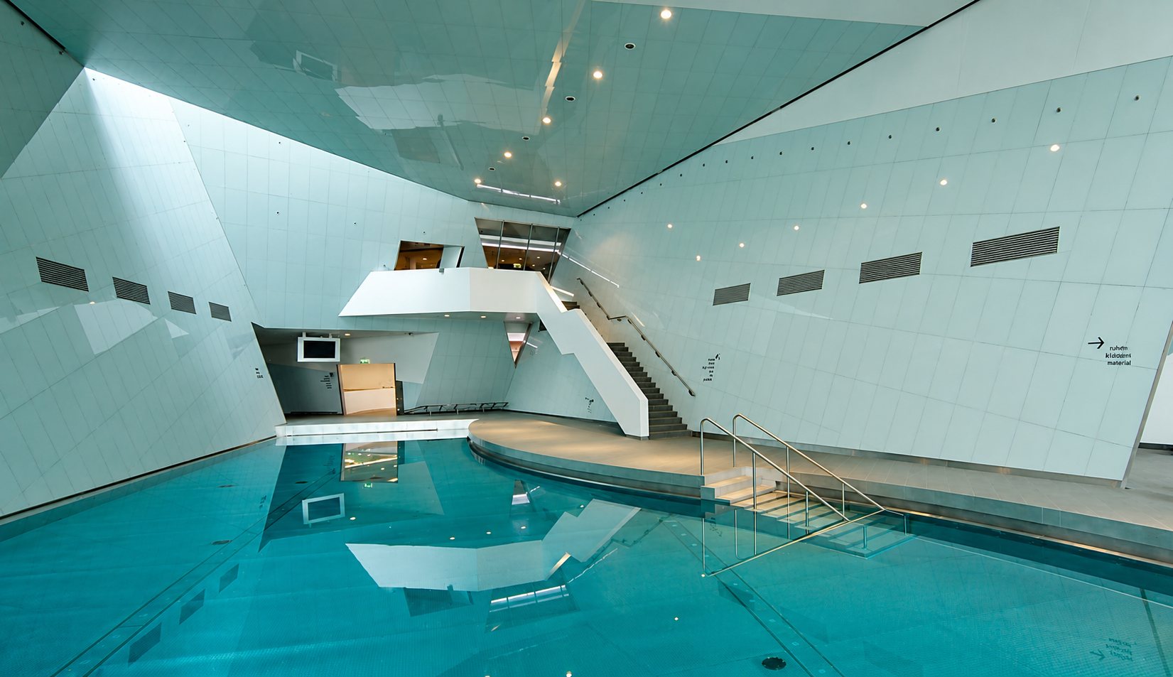 Moderne Schwimmhalle mit schräg verlaufenden, hell gefliesten Wänden, türkisfarbenem Becken und einer Treppe mit Edelstahlgeländer